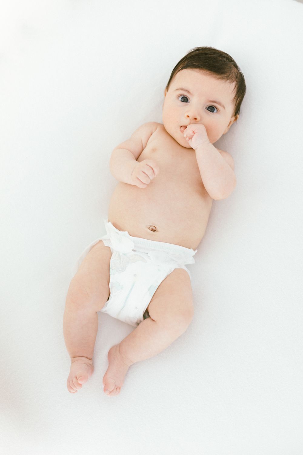 Baby wearing white diaper lying on back against white backdrop.