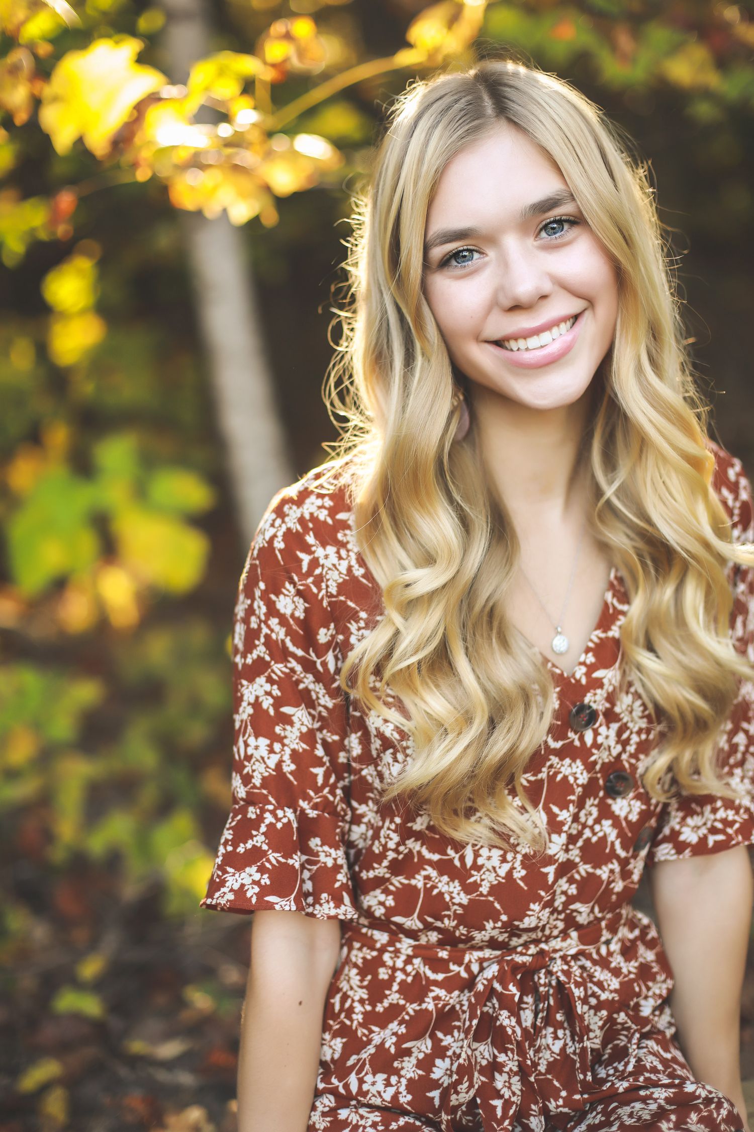 Woman in floral dress poses outdoors with autumn foliage in background.