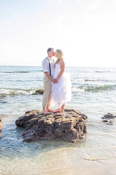 A romantic beach wedding portrait with couple standing on ocean rocks at sunset.