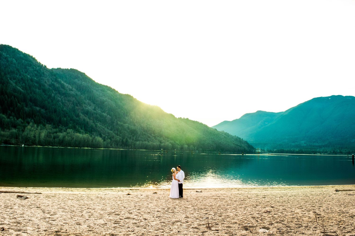 A couple stands on a beach with mountains and a serene lake in the background at sunset.