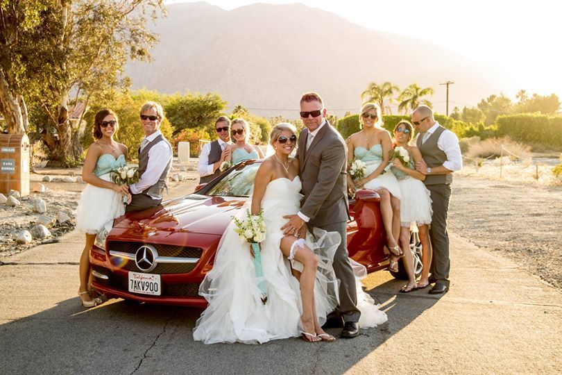 A wedding party poses playfully around a red car at sunset.
