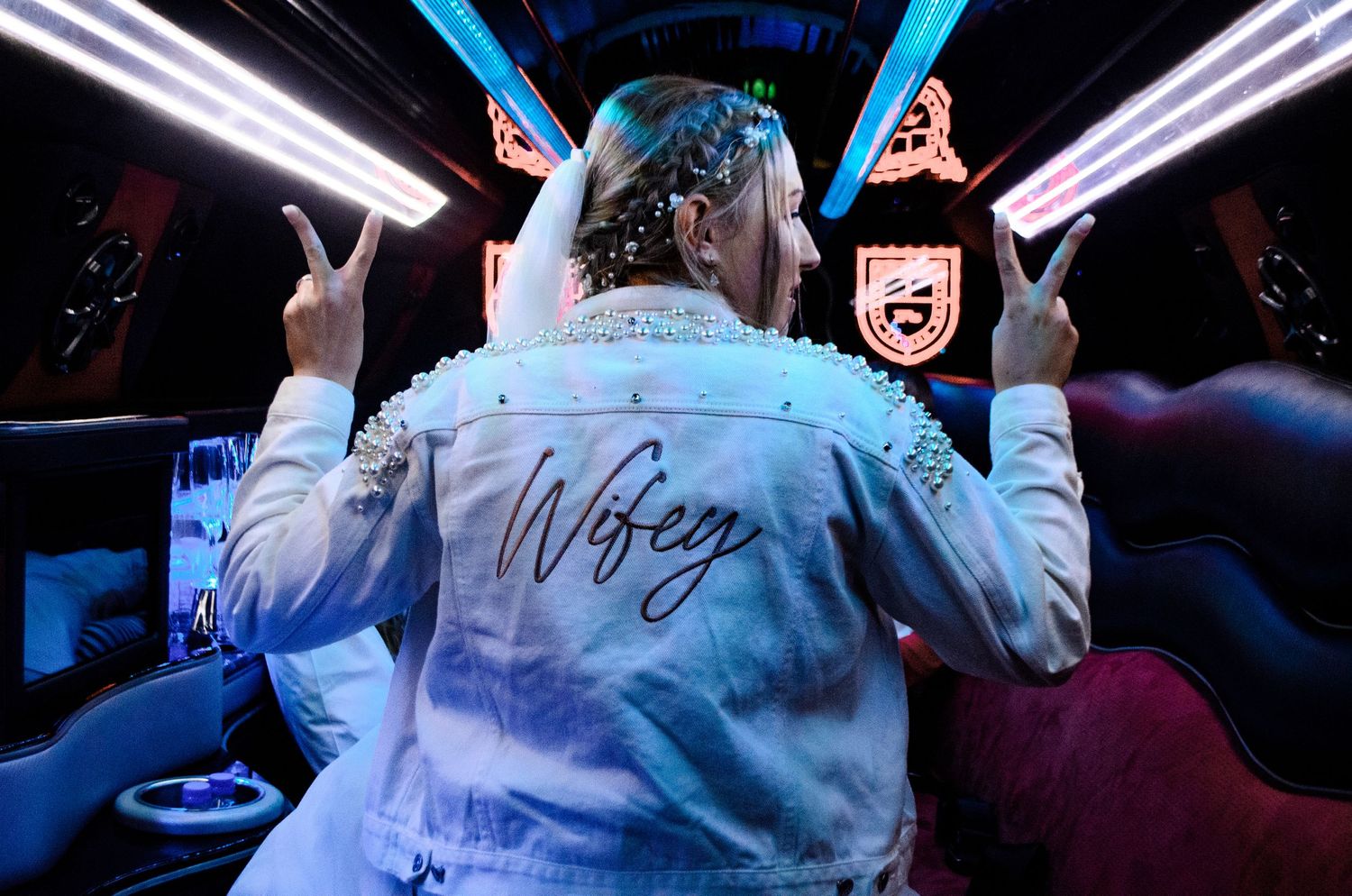 Person wearing a white jacket with 'Wifey' written on it sits in a party bus with neon lights.