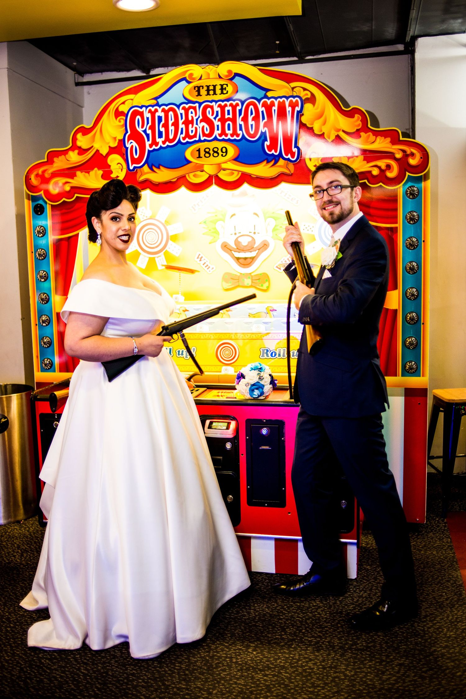 Wedding couple poses playfully with prop guns at a vintage sideshow arcade game display.