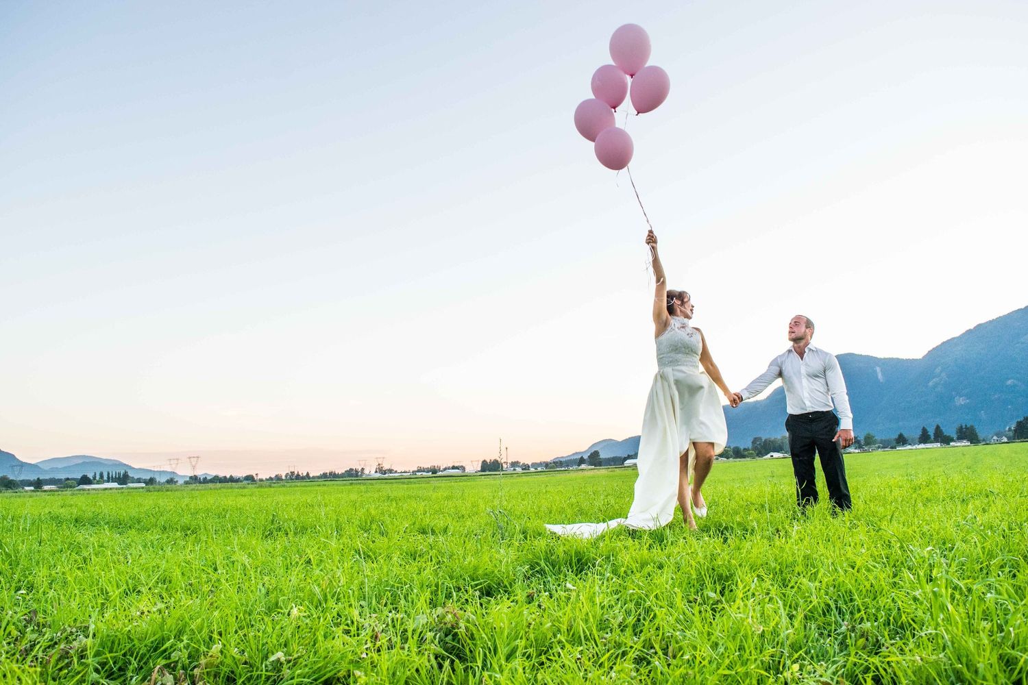 A couple holds pink balloons while standing in a green meadow with mountains in the background.