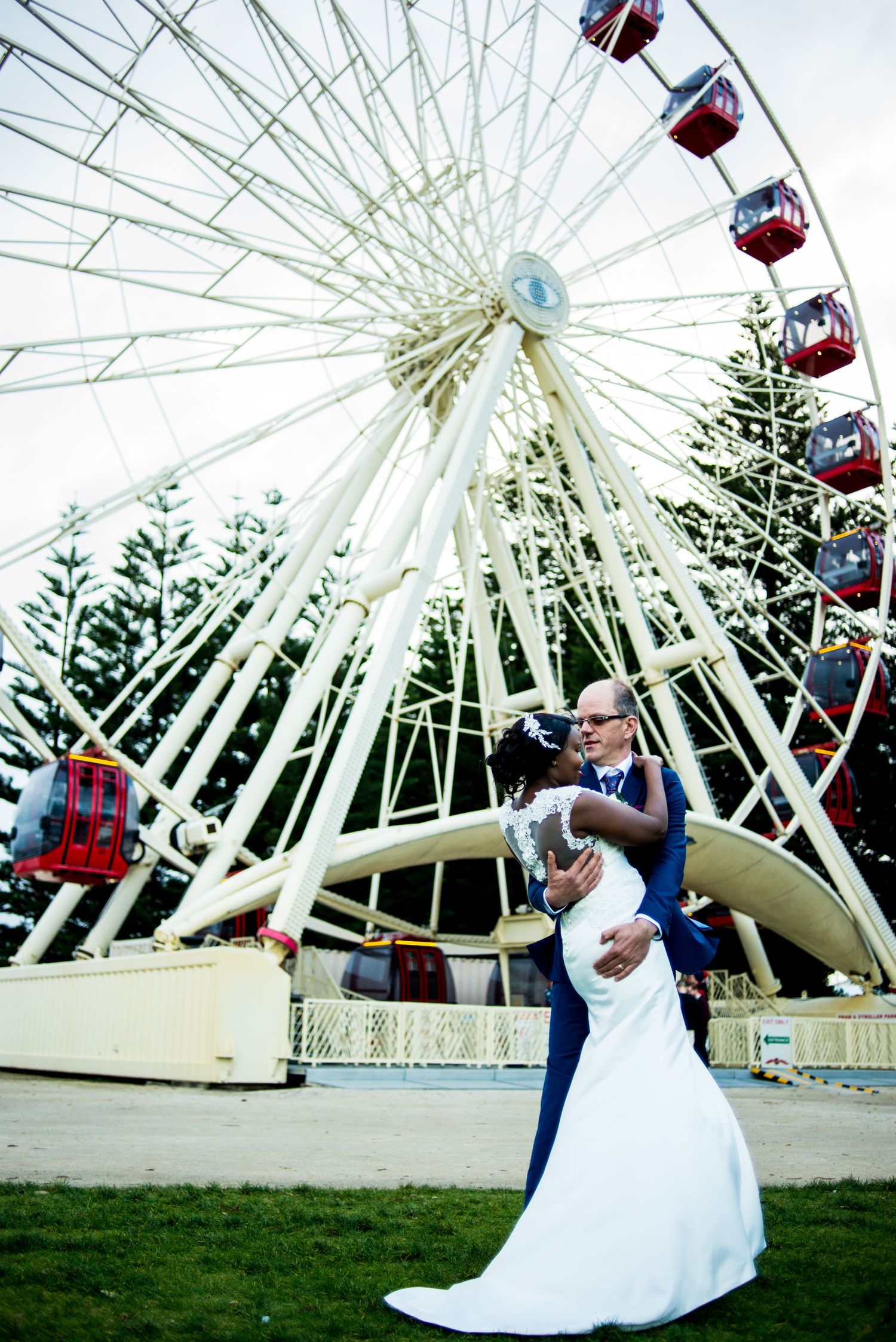 Wedding couple poses dramatically in front of a towering white Ferris wheel.