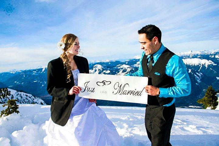 A couple in winter attire holds a 'Just Married' sign on a snowy mountain peak.