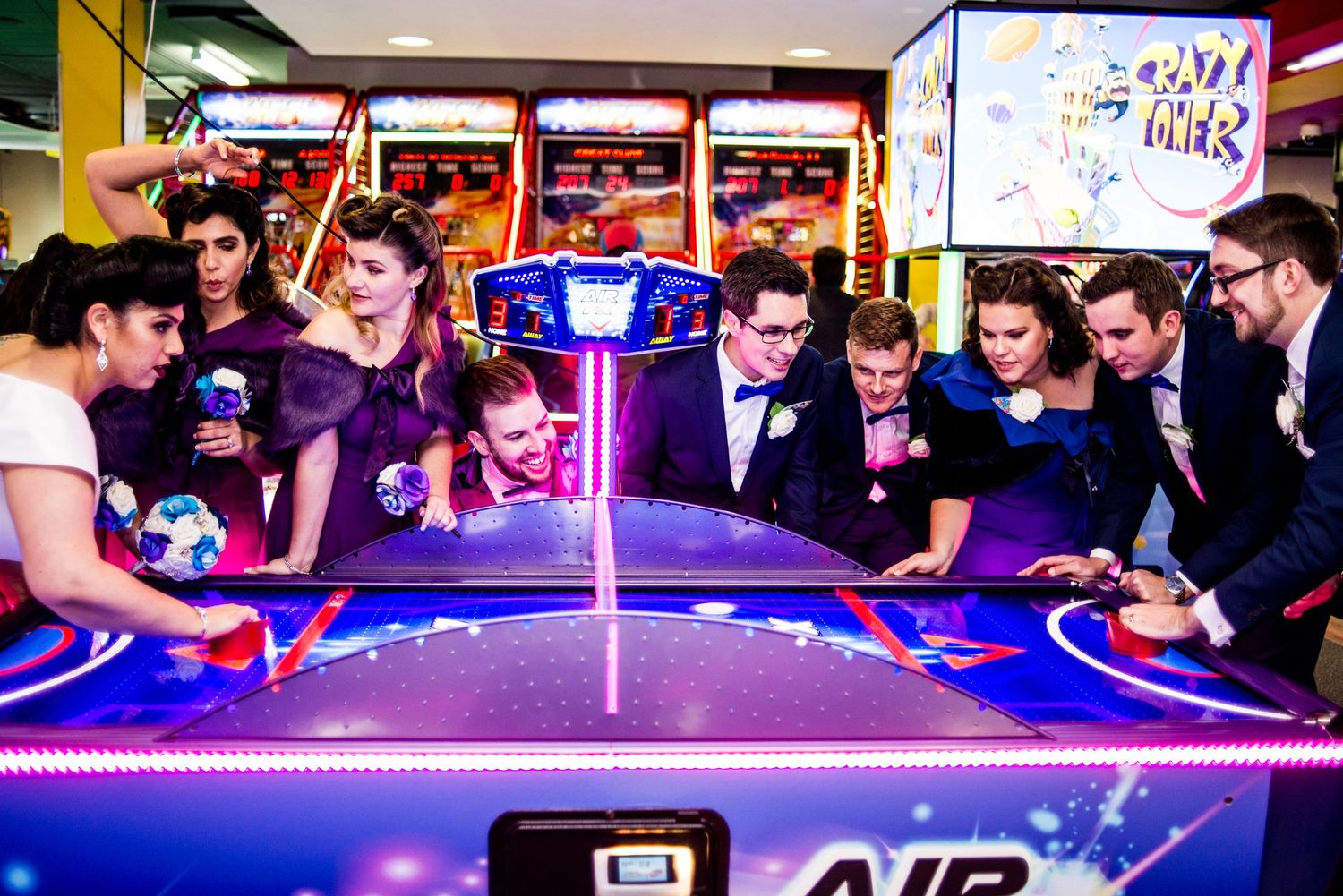 Wedding party gathered around an illuminated air hockey table in an arcade setting.