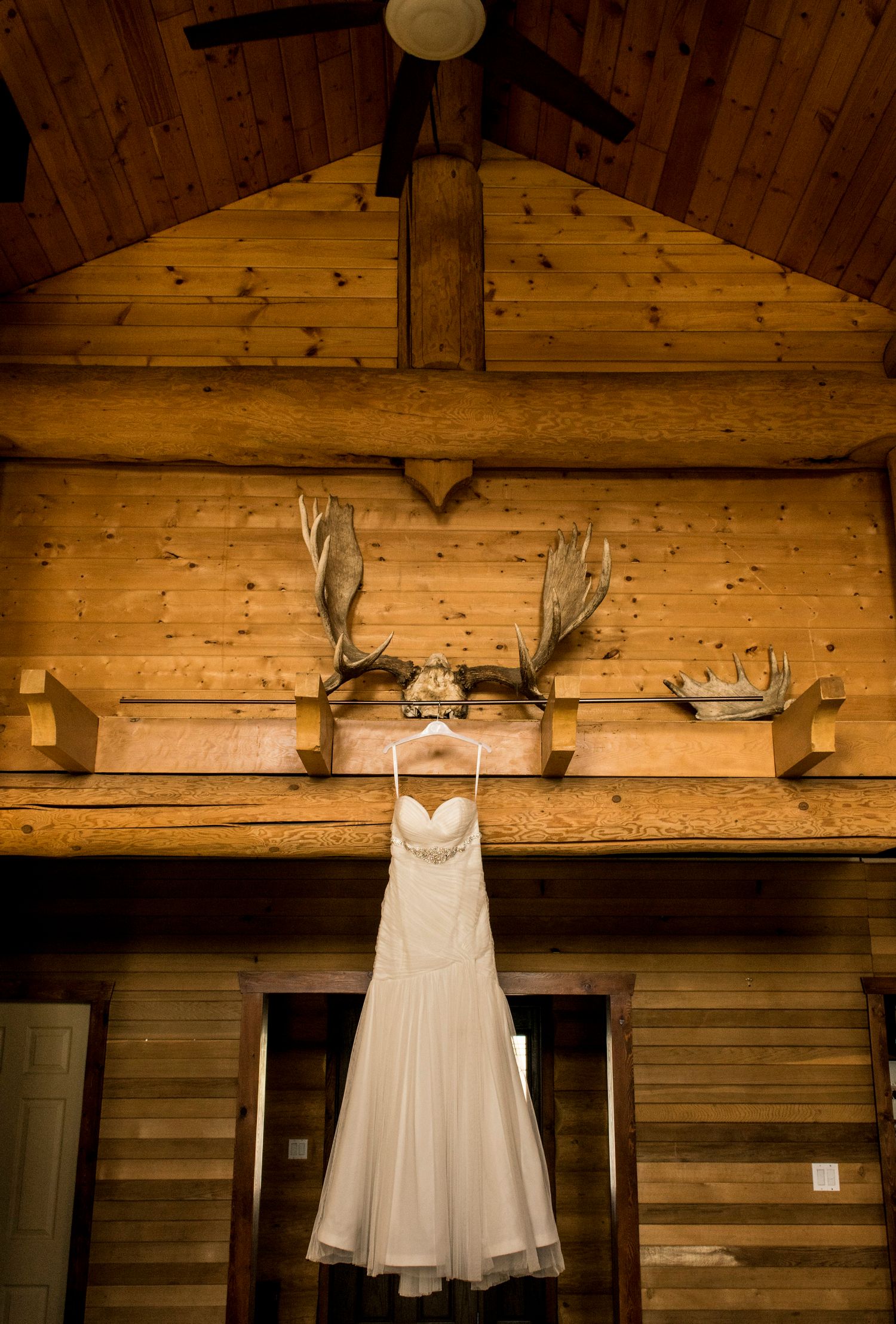 A white wedding dress hangs from antler mounts on a rustic wooden wall.