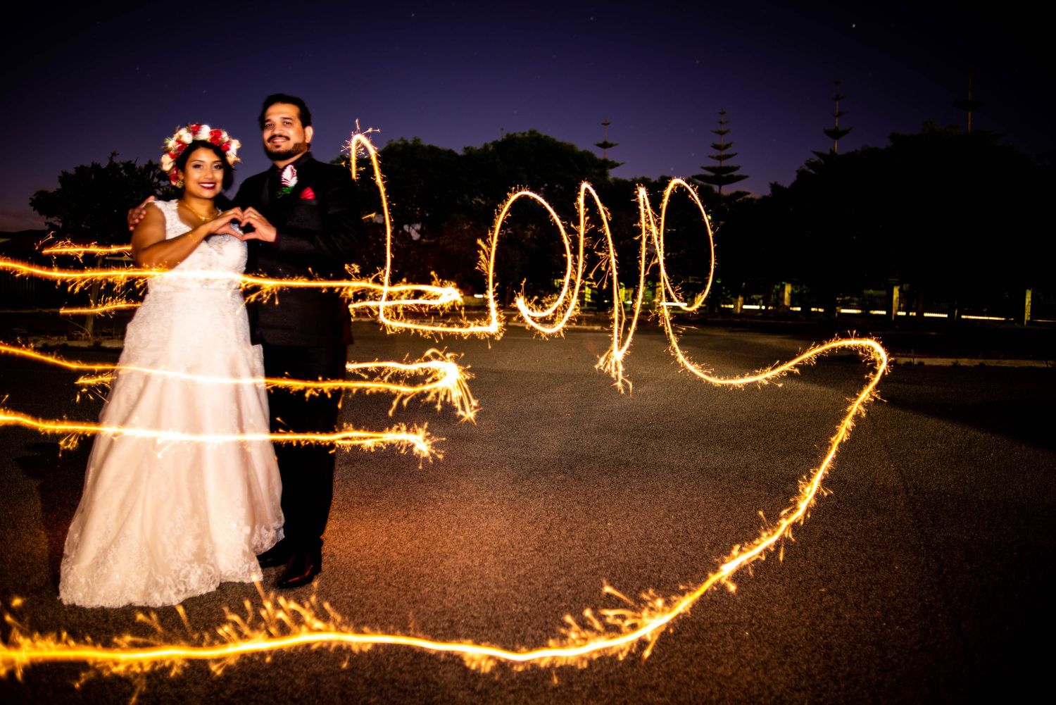 Light painting with sparklers creates swirling patterns around a couple in wedding attire at night.