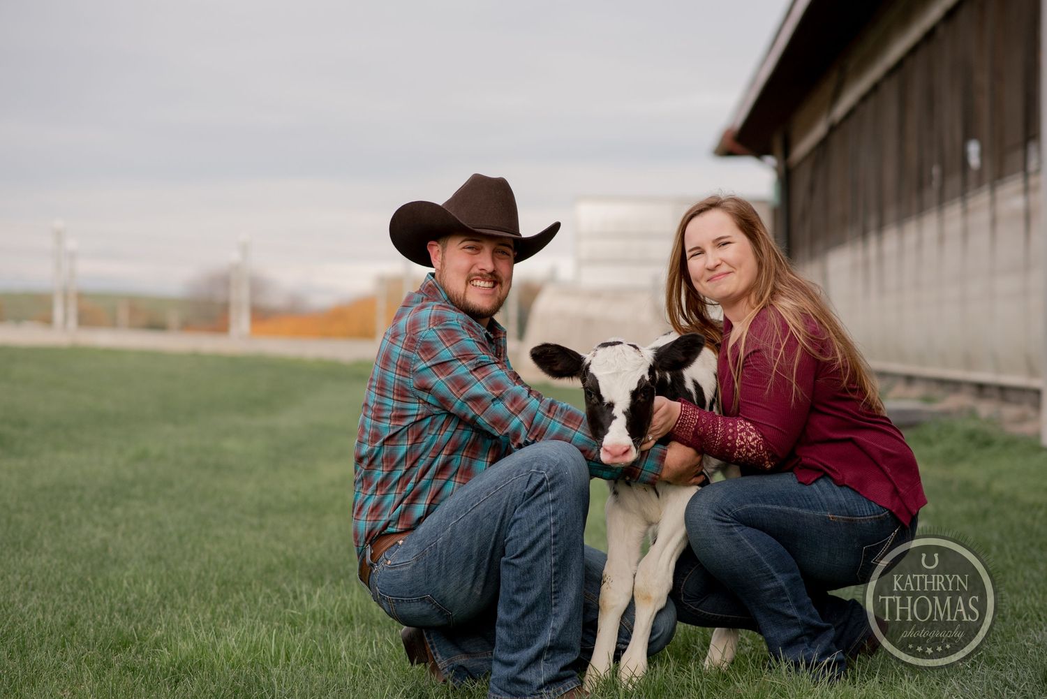 Couple poses with young Holstein calf on grass outside dairy barn.