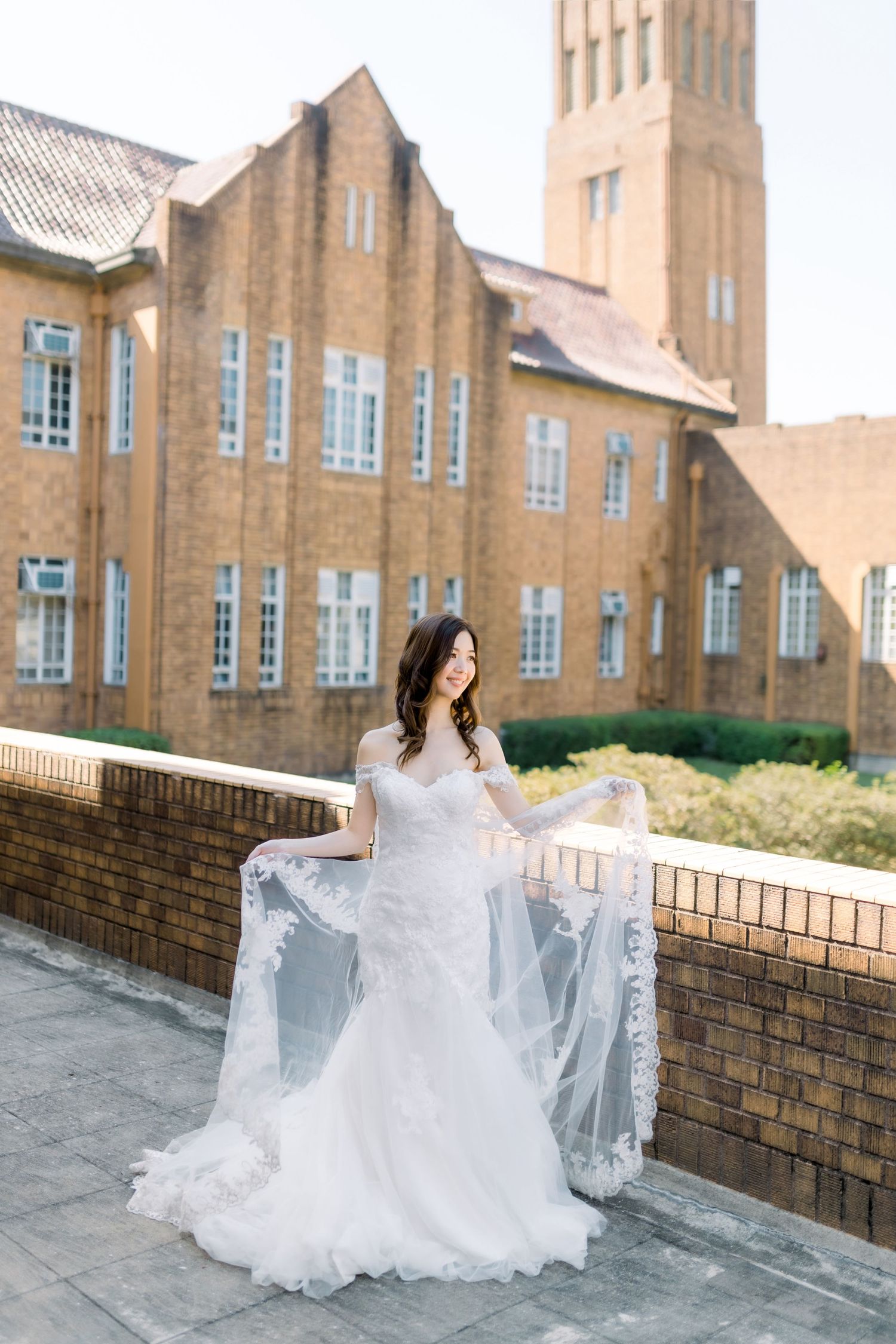 Elegant bride in lace gown poses against historic brick building with tall tower.