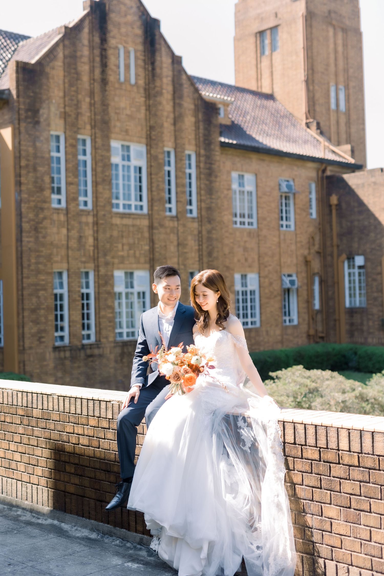 Wedding couple sits on brick wall in front of historic church building.