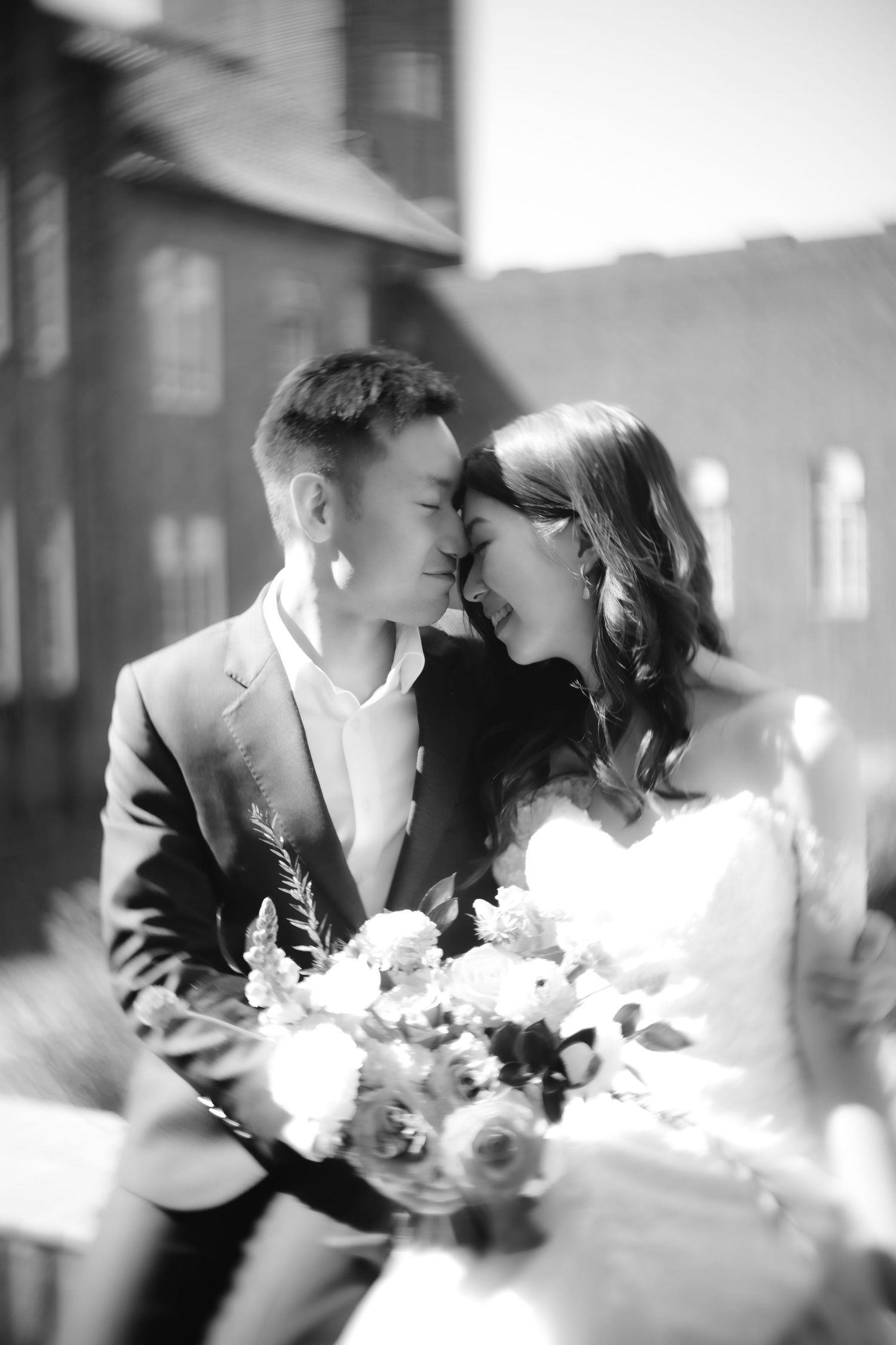 An artistic black and white photo of a couple with a wedding bouquet against a building backdrop.