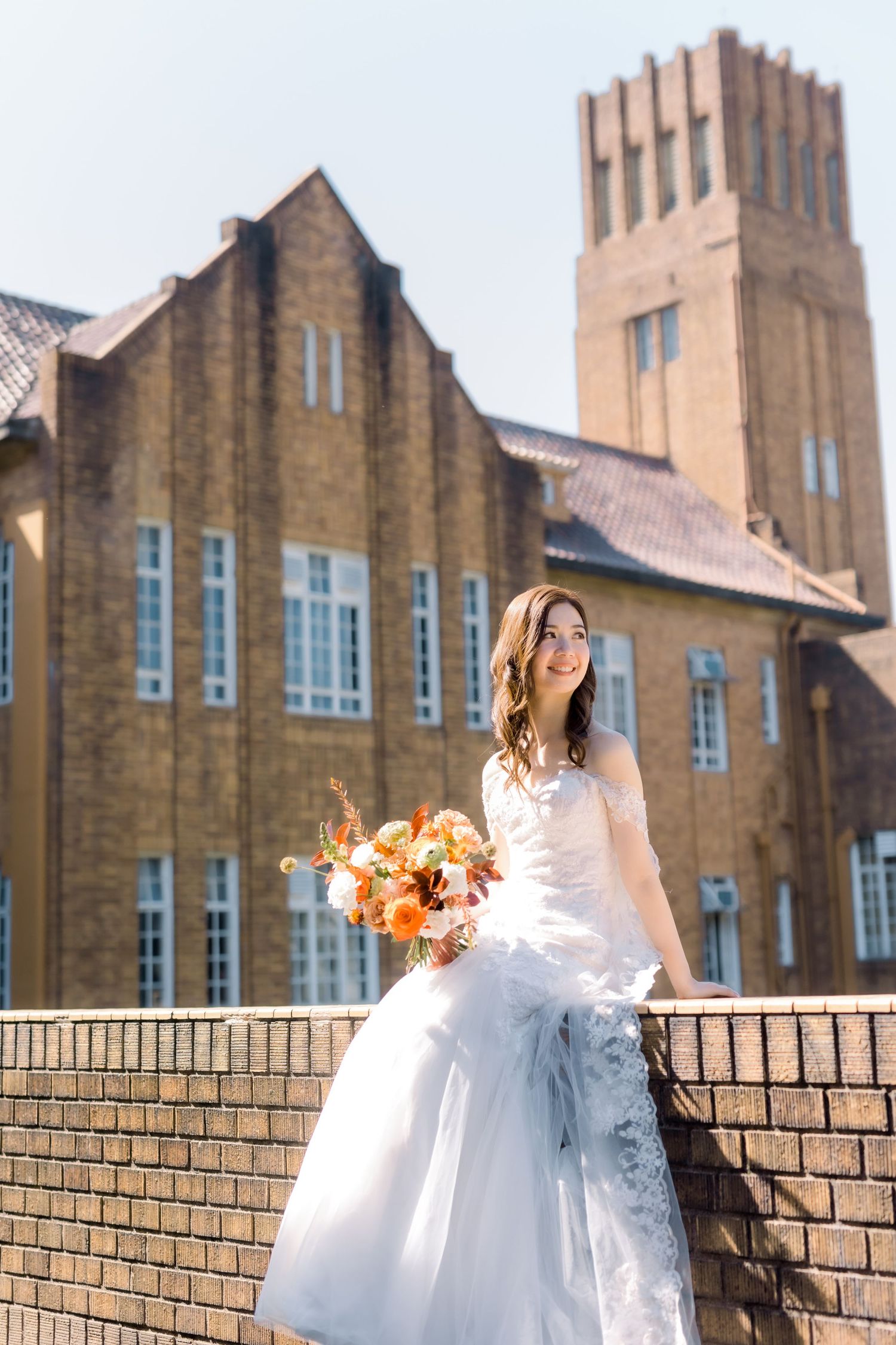 A bride in white holds orange flowers while posing against a brick church with a tower.