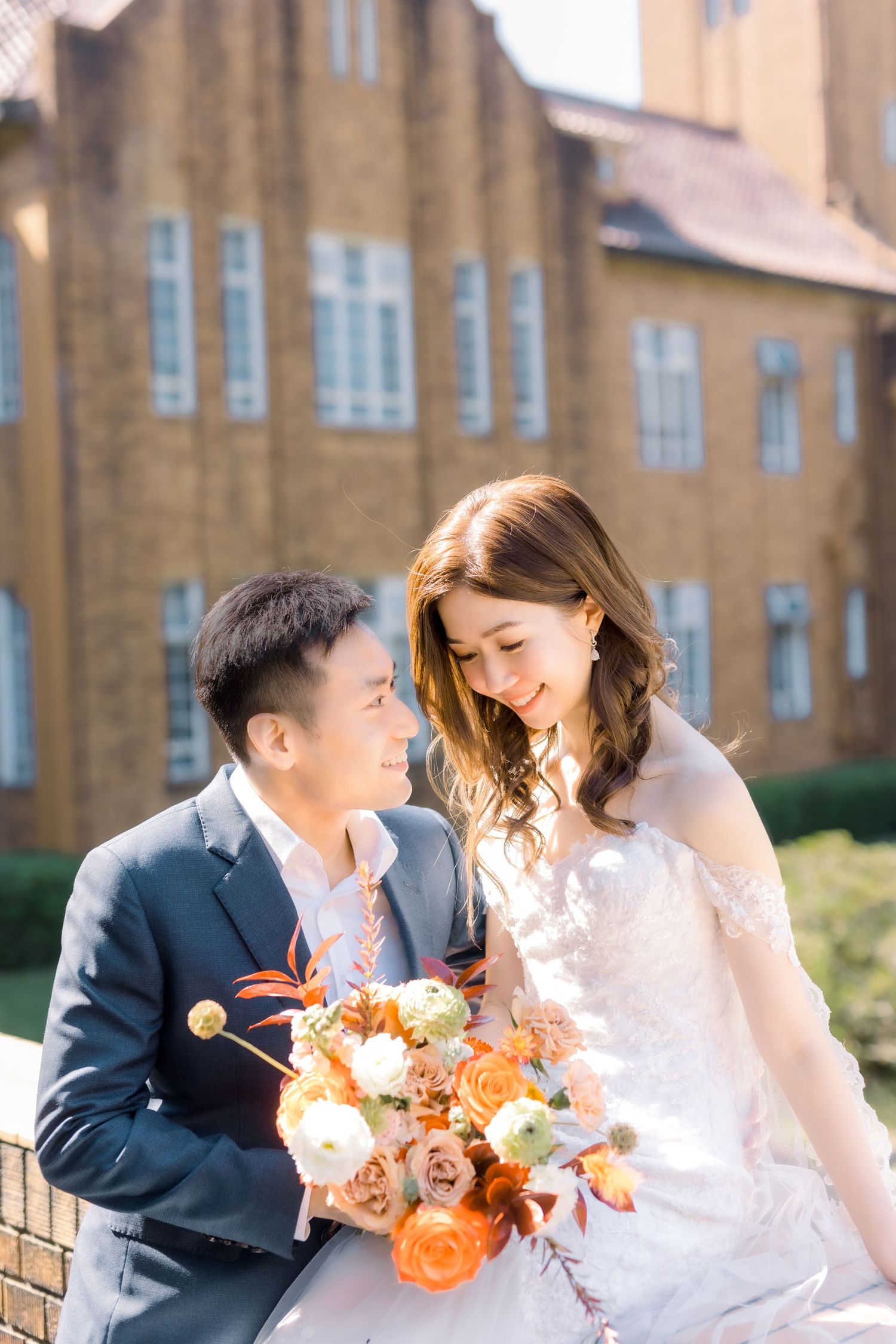 A couple shares a joyful moment with orange flowers against a historic brick building.