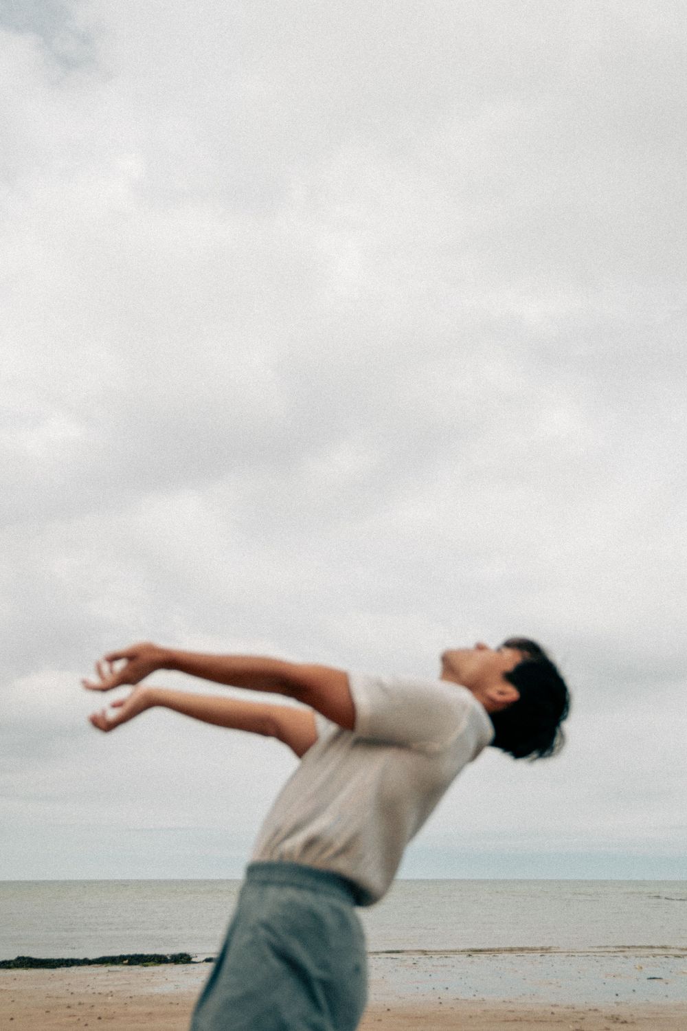 A figure arches backward in an expressive dance move against a cloudy sky at the beach.