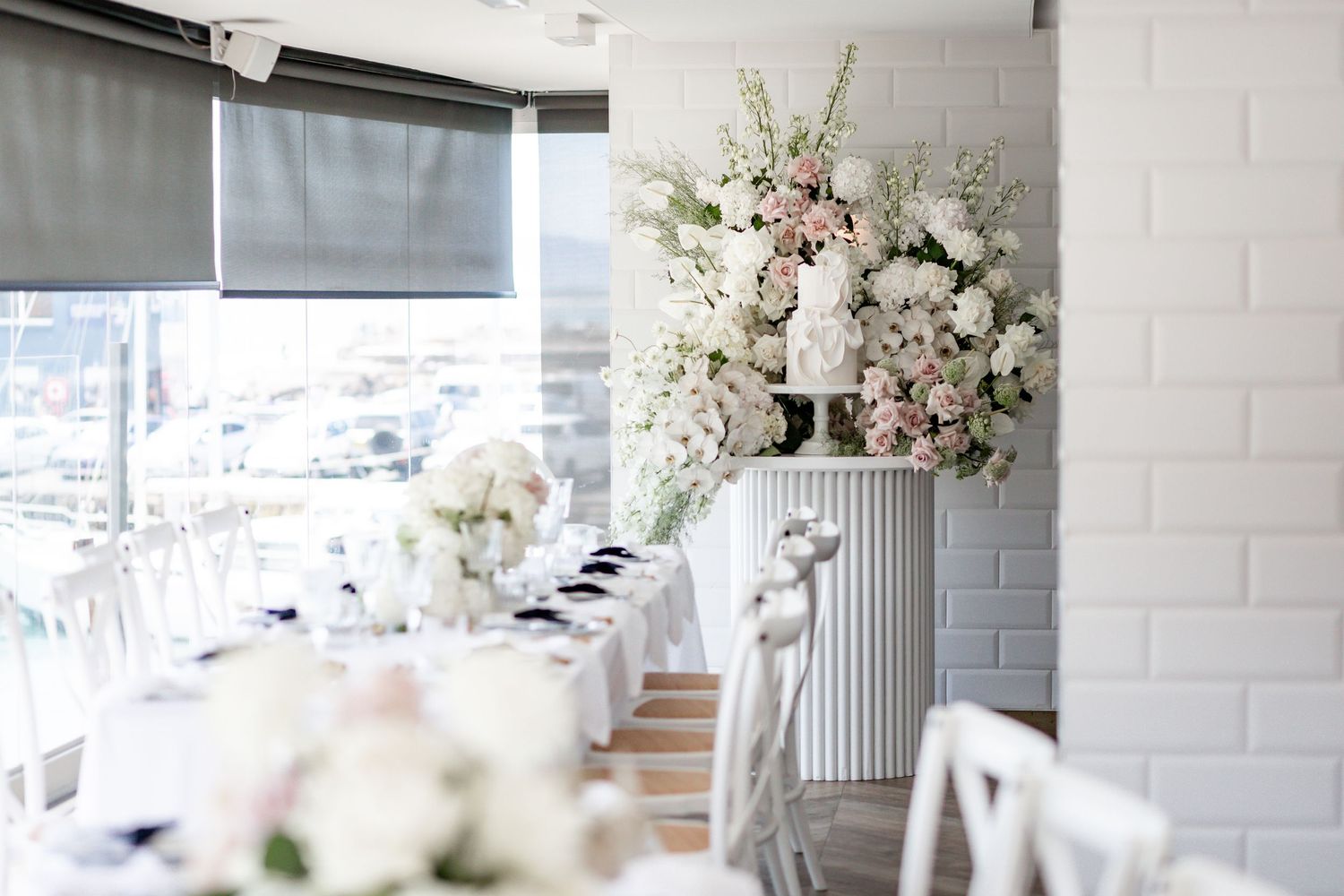 White and blush floral arrangement on a classical column against white brick wall in an event space.