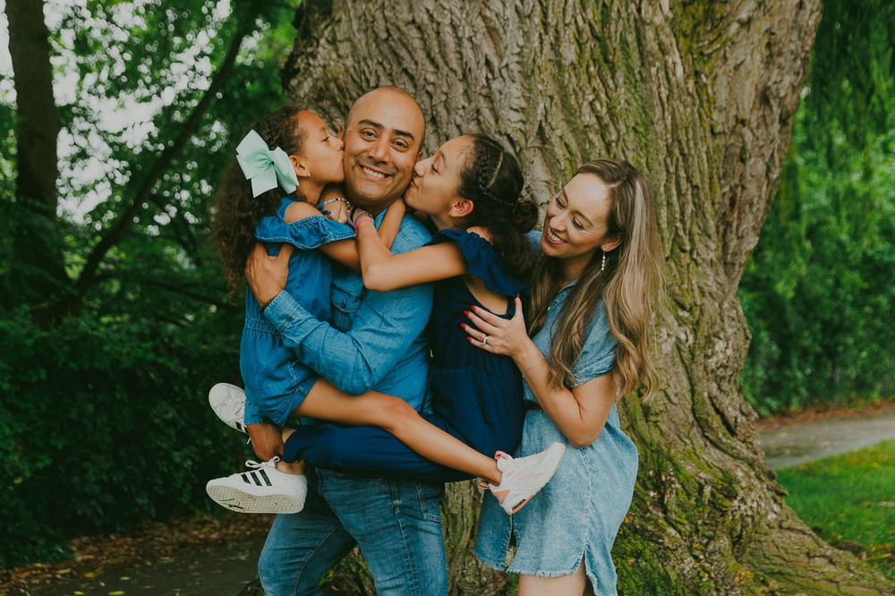 A joyful family moment captured against a large tree trunk in a park setting with coordinated blue outfits.