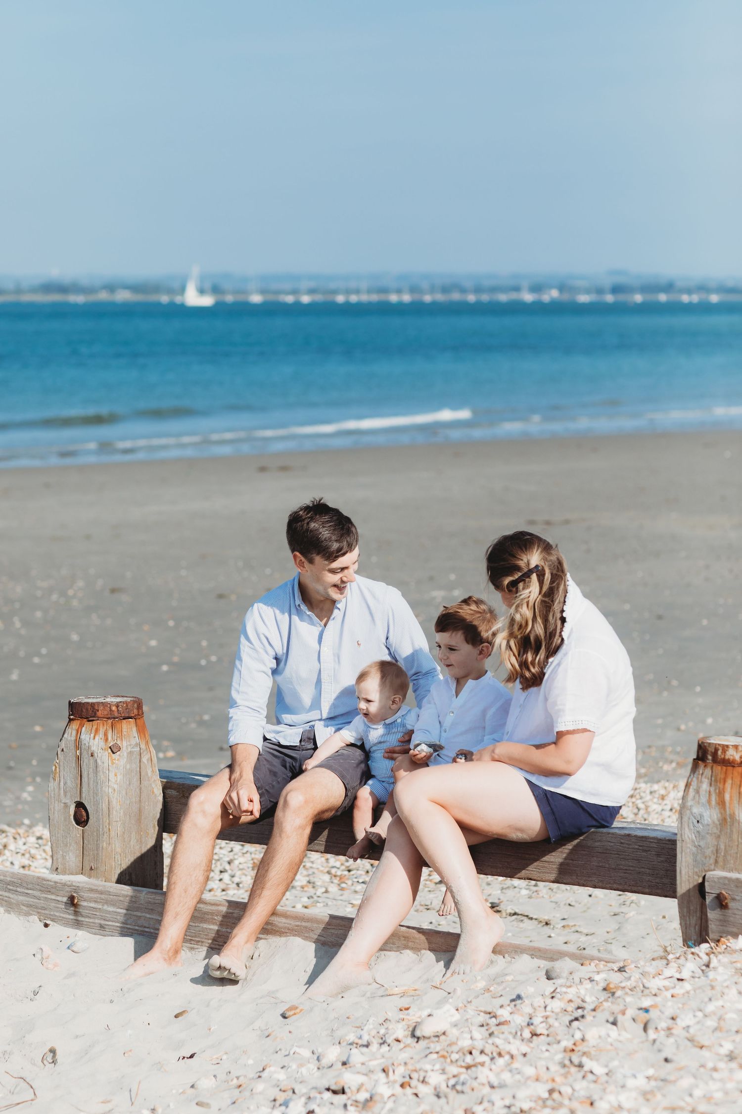 A family relaxes together on wooden posts at the beach with turquoise water and a sailboat in the background during a family beach photoshoo