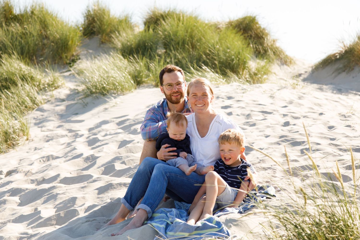 Family enjoying a relaxed moment together on sandy beach dunes surrounded by grass during a family beach photoshoot