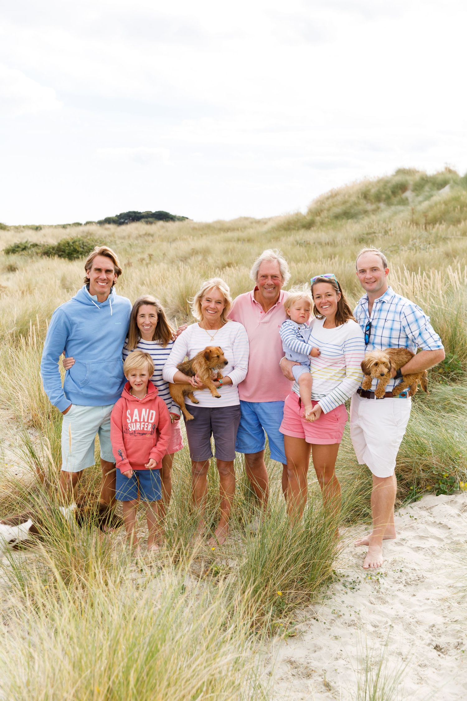 Multi-generational family with dogs posing together in beach grass dunes on a sunny day during a family beach photoshoot