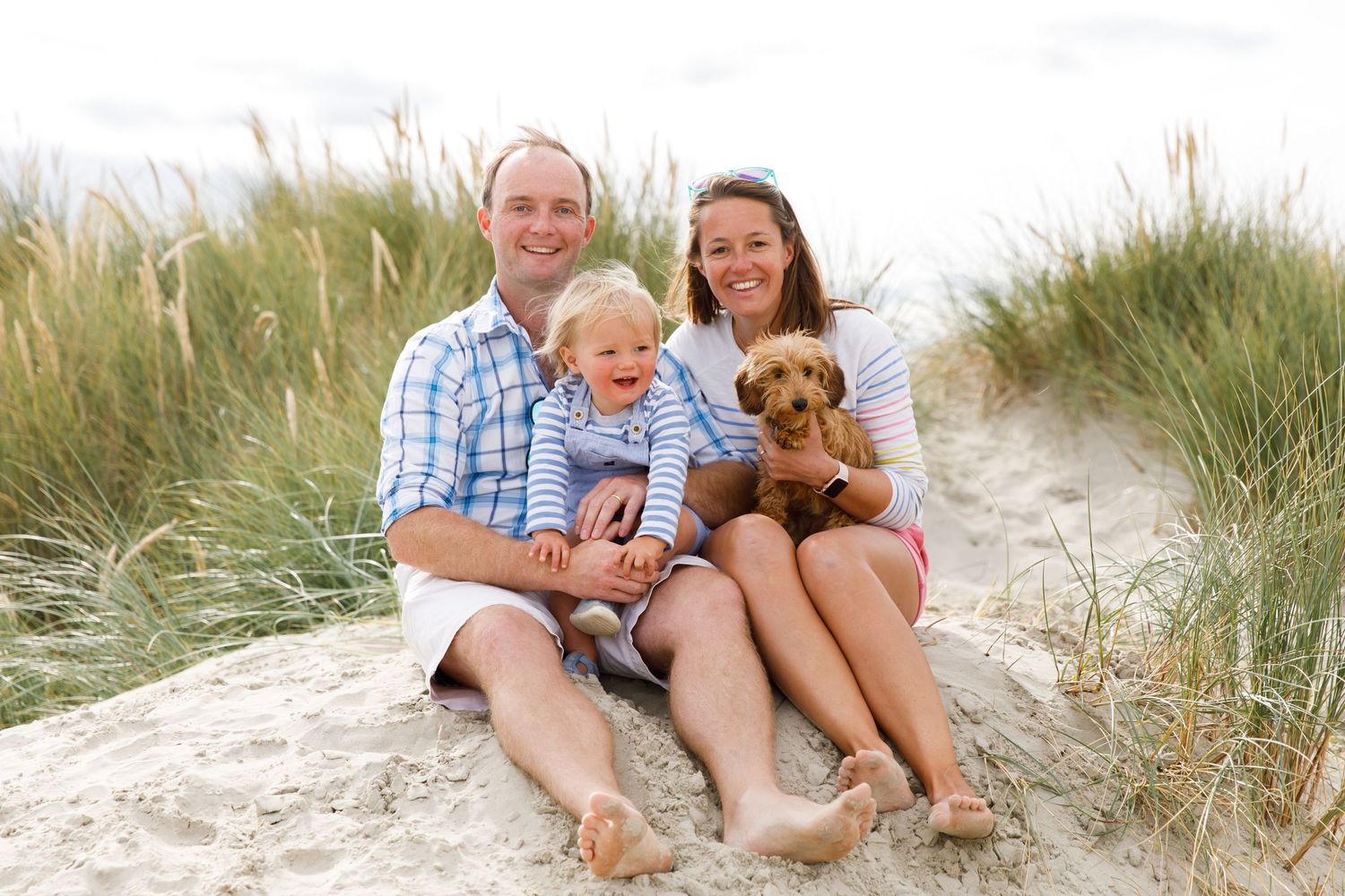 A couple sits on beach dunes with their small dog enjoying a sunny day together during a family beach photoshoot