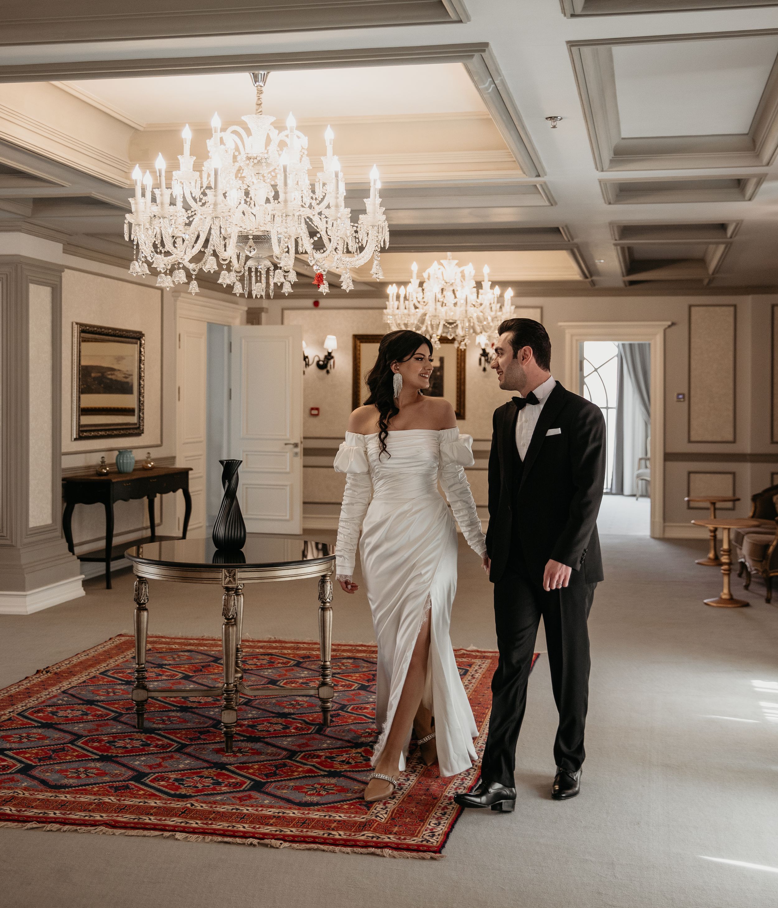 Elegant couple in formal attire walking under crystal chandeliers in a luxurious room.
