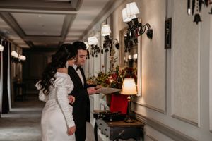 Couple in formal attire admiring elegant hallway decor with antique wall sconces and floral arrangements.