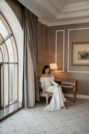 Bride in off-shoulder gown seated elegantly by arched window in luxury hotel room.