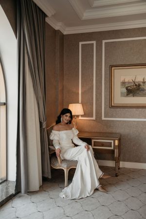 Elegant bridal portrait in luxurious hotel room with dramatic window light and classic furniture.