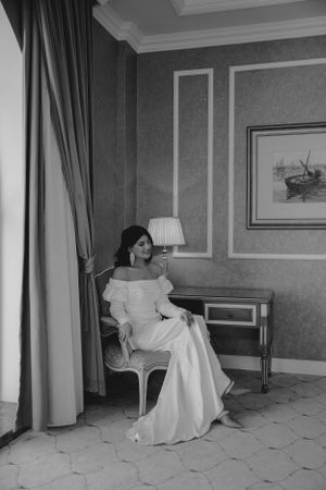 Black and white portrait of bride seated by window in elegant hotel room with classic decor.