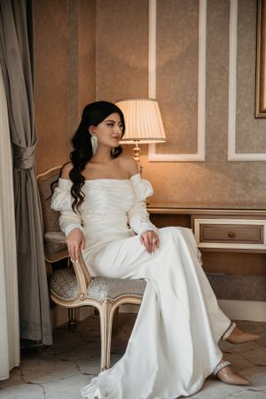 Bride in off-shoulder gown poses gracefully on vintage chair in elegant hotel setting.