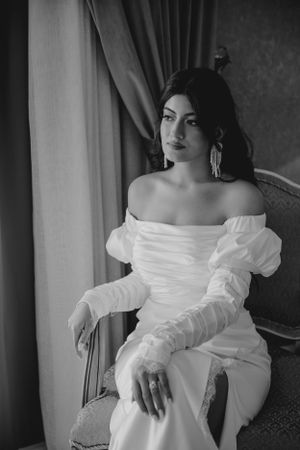 Elegant off-shoulder white dress with ruched sleeves photographed in black and white against a window and curtain.
