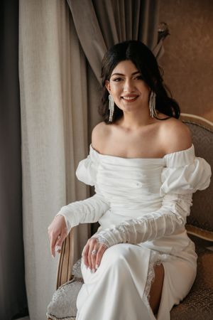 Modern bride in form-fitting white dress with dramatic puff sleeves poses in vintage chair.