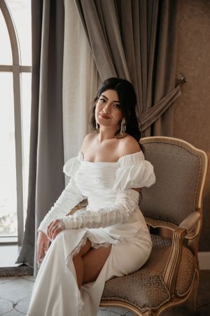 Bride in elegant white dress with puff sleeves sits in vintage chair by window.