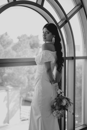 Bride in off-shoulder wedding dress holding bouquet poses by arched window.