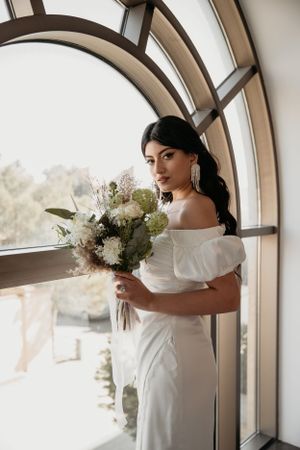 Elegant bridal pose with white floral bouquet framed by architectural arched window.