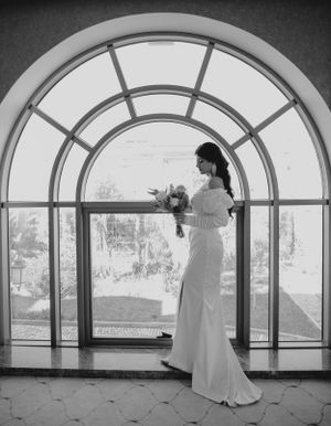 A black and white wedding photo featuring an elegant bride in a fitted gown standing in front of an arched window.