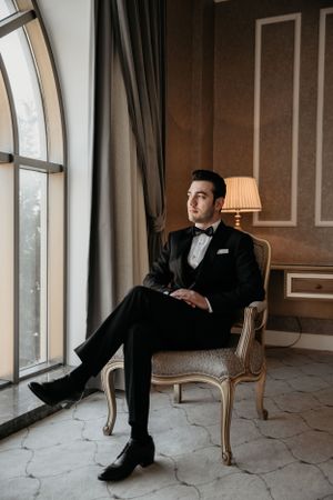 Elegant groom in classic tuxedo seated in vintage chair by large arched window.