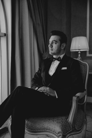 Black and white portrait of groom in tuxedo seated in vintage chair with dramatic window lighting.