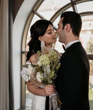 Intimate wedding moment captured near arched window with delicate bouquet and formal attire.
