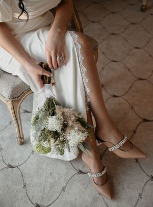 Detail shot of bride's embellished shoes and flowing dress with dried flower bouquet on tiled floor.