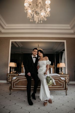 A newlywed couple poses in formal attire beneath a crystal chandelier in an elegant hotel suite.