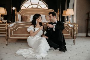 A couple in wedding attire sharing an intimate moment while seated on the floor near an ornate bed.