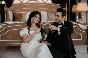 A playful moment captured of a wedding couple sharing drinks together in a luxurious bedroom setting.