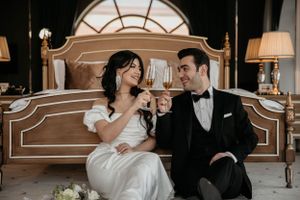 A romantic wedding photo of a couple toasting with champagne while sitting by an elegant headboard.