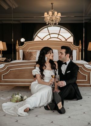A candid wedding photo showing a couple sharing drinks while seated on the floor in an elegantly lit room.