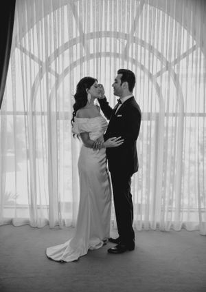 A black and white romantic portrait of a couple in wedding attire against sheer curtains with arched window.