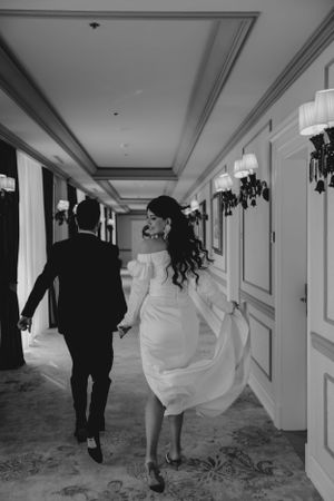 A couple running playfully down an elegant hotel hallway with white walls and ornate light fixtures.