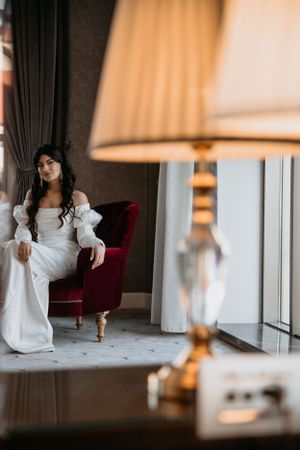 An elegant scene with a person in a white dress seated on a red velvet chair near a window.